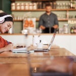 woman working on a laptop in a cafe with a man at the counter in the background